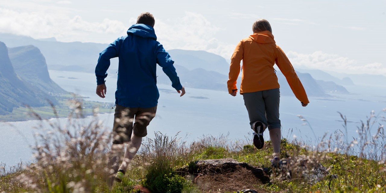Man en vrouw met softshell jassen rennend op een berg met uitzicht op landschap